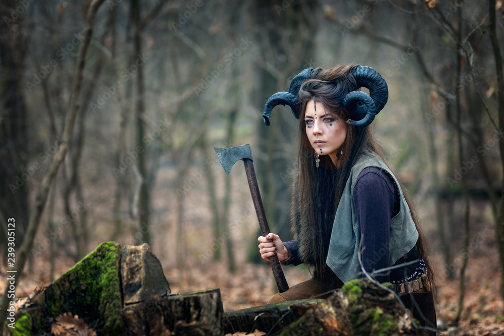 Beautiful young woman warrior with blue eyes wearing ram horns and specific  makeup holding ax hiding behind stumps in forest, stalking and ready to  attack or defend her land. Stock 写真