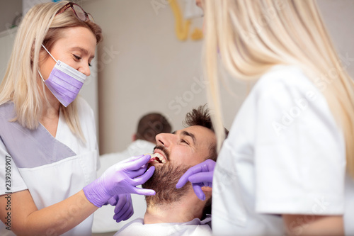 Two young female dentists working in dental clinic. Whitening male patient s teeth and using tone chart to mach color. Health care and medicine concept.