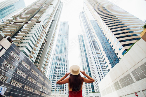 Photography Back view of young woman in red summer dress holding with both hands straw hat looking up on skycrapers at downtown in modern city