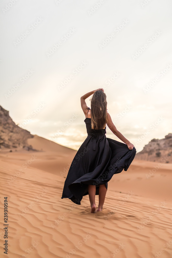 Rear view of young woman in black dress dancing in sandy desert at sunset Rear view of young woman in black dress dancing in sandy desert at sunset