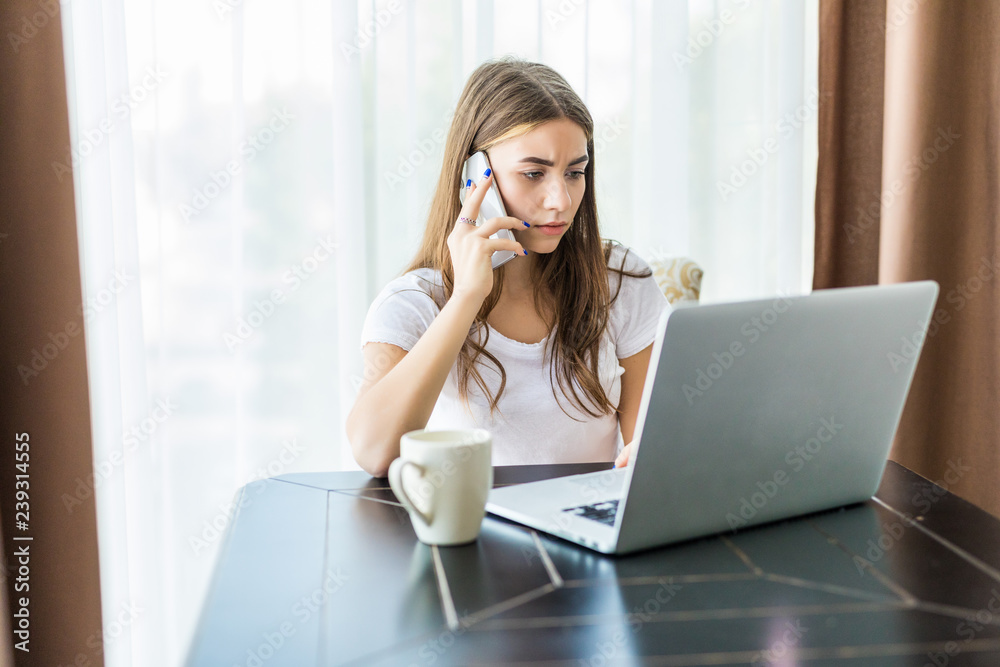 Cute young woman talk phone while working on laptop computer at desk next to coffee cup at home with large window in background Cute young woman talk phone while working on laptop computer at desk next to coffee cup at home with large window in background