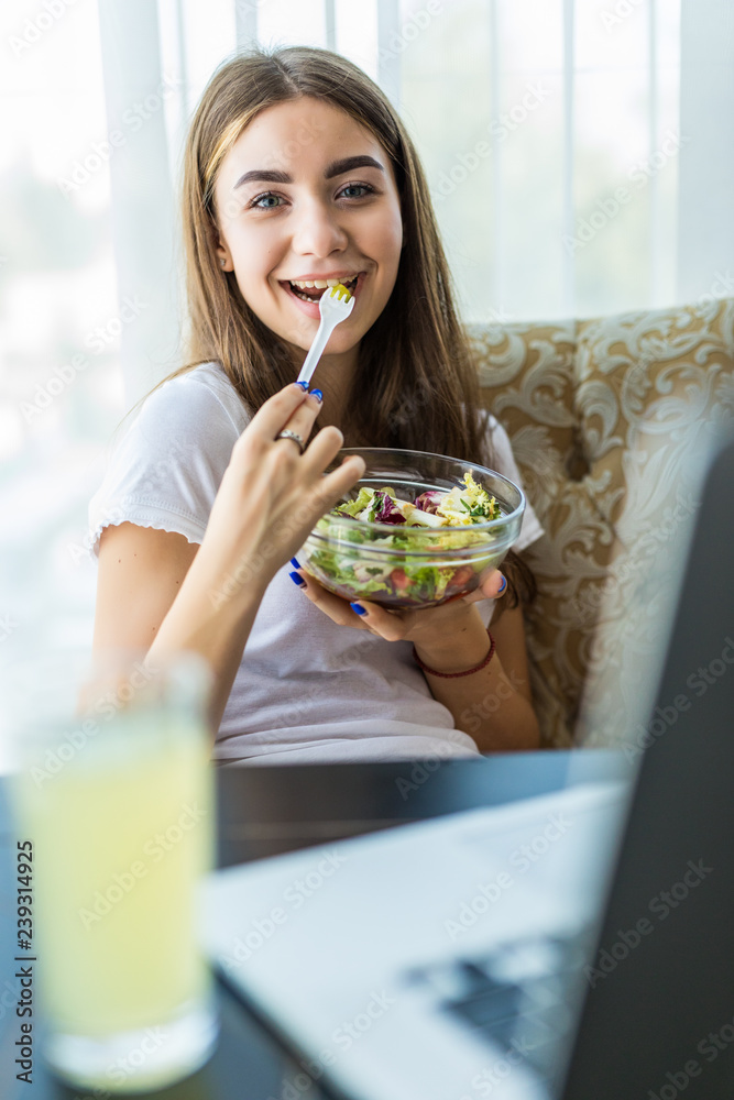 Portrait of a happy young woman eating fresh salad from a bowl while sitting and using laptop computer at home Portrait of a happy young woman eating fresh salad from a bowl while sitting and using laptop computer at home