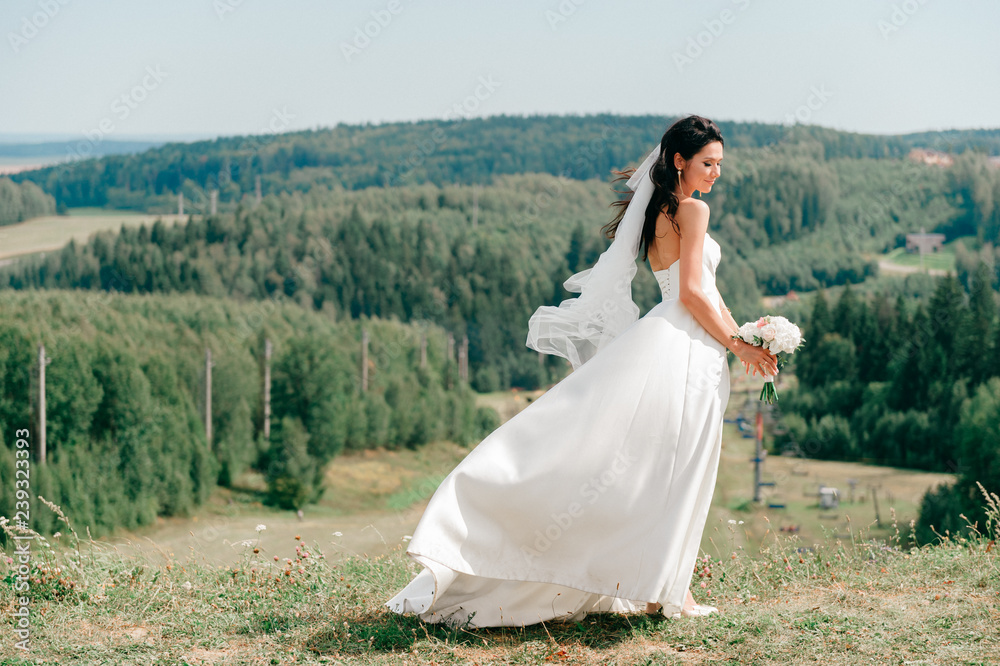 Happy brunette bride in beautiful white dress posing with boquet of flowers on top of mountain  with fabulous landscape view. Bridal outdoor photosession. Cheerful brunette bride lifestyle portrait.