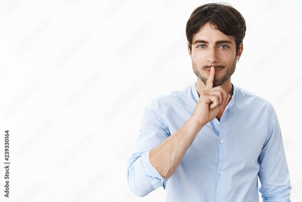 Waist-up shot of sexy handsome successful man with beard and blue eyes smiling naughty making shush gesture over folded lips asking keep voice down as preparing secret surprise over grey wall Waist-up shot of sexy handsome successful man with beard and blue eyes smiling naughty making shush gesture over folded lips asking keep voice down as preparing secret surprise over grey wall