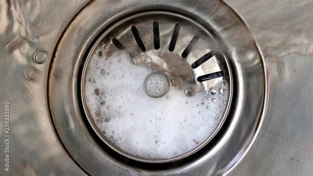 Water with soap bubbles flows into the drain hole in the metal sink, macro view. Mechanically adjustable drain plug closeup. White foam with bubbles of cleaner in a washbasin. Water with soap bubbles flows into the drain hole in the metal sink, macro view. Mechanically adjustable drain plug closeup. White foam with bubbles of cleaner in a washbasin.