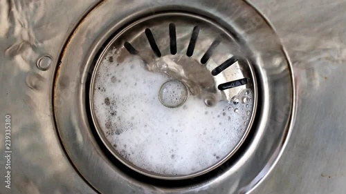 Water with soap bubbles flows into the drain hole in the metal sink, macro view. Mechanically adjustable drain plug closeup. White foam with bubbles of cleaner in a washbasin. photo