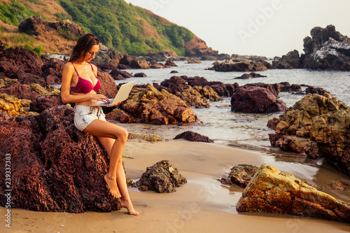 Lady freelancer in red bikini swimsuit with a laptop remote work on the beach on the sunset.freelancing girl working near the rocks and the sea.business women on vacation in a tropical desert paradise