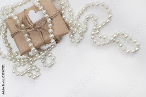 Gift with heart in craft paper and pearl necklace on the white background. Romance, love concept. Top view, flat lay, copy space, card layout