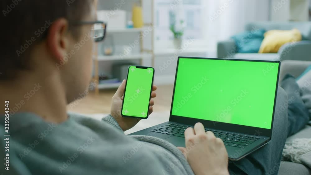Young Man at Home Works on a Laptop Computer with Green Mock-up Screen, while Holding Smartphone with Chroma Key Display. He's Sitting On a Couch in His Cozy Living Room. Over the Shoulder Camera Shot Young Man at Home Works on a Laptop Computer with Green Mock-up Screen, while Holding Smartphone with Chroma Key Display. He's Sitting On a Couch in His Cozy Living Room. Over the Shoulder Camera Shot