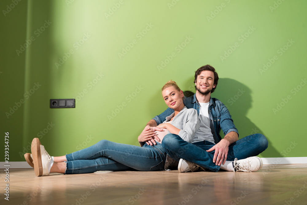 smiling man and pregnant wife sitting on floor and looking at camera smiling man and pregnant wife sitting on floor and looking at camera