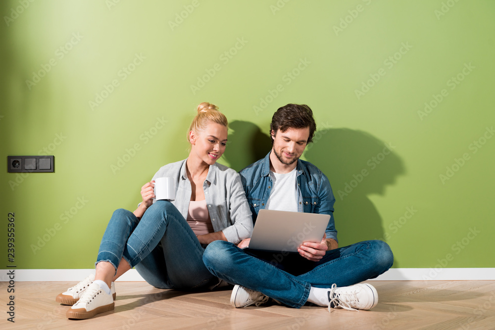 handsome man and attractive girl with coffee cup sitting on floor and looking at laptop screen handsome man and attractive girl with coffee cup sitting on floor and looking at laptop screen