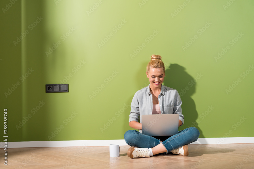 smiling girl typing on laptop keyboard and sitting on floor by green wall smiling girl typing on laptop keyboard and sitting on floor by green wall