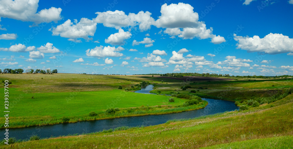 Sunny summer scene with river curve,fields,green hills,distant woods and beautiful clouds in blue sky.