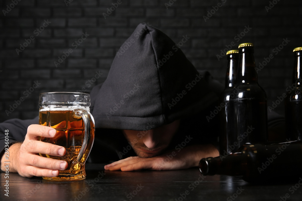 Drunk young man with beer at table near black brick wall. Alcoholism concept