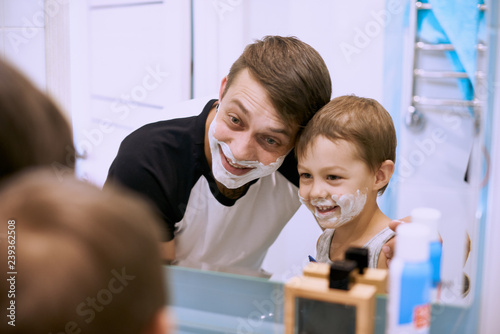 Man and little boy with shaving foam on their faces looking into the bathroom mirror and laughing. Father and son having fun while shaving in bathroom.