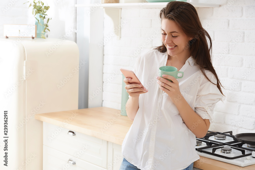 Young woman using smartphone leaning at kitchen table with coffee mug and organizer in a modern home. Smiling woman reading phone message. Brunette happy girl typing a text message. Young woman using smartphone leaning at kitchen table with coffee mug and organizer in a modern home. Smiling woman reading phone message. Brunette happy girl typing a text message.