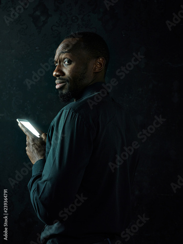 Studio shot of young serious black African man thinking while talking on mobile phone against black studio background