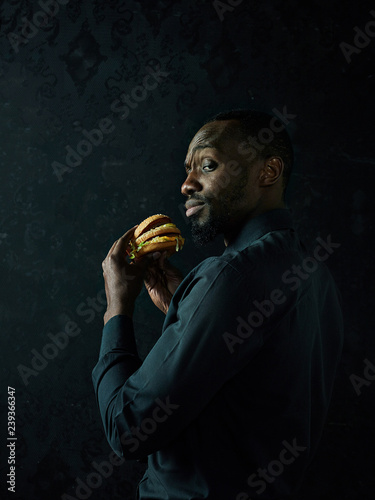 Fototapeta The young african american man eating hamburger and looking away on black studio
