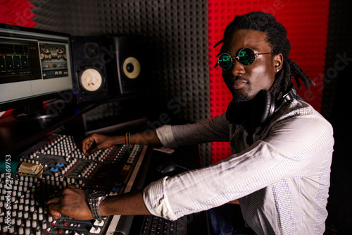 African man sound engineer with headphones at work sitting mixing console. Side view © pantovich