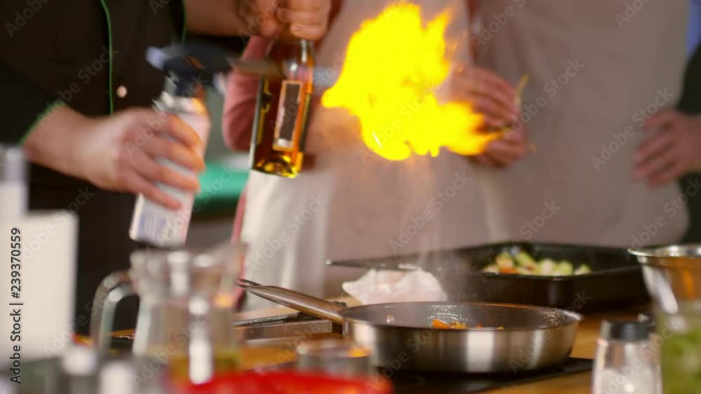 Medium shot of male chef pouring alcohol on frying vegetables, setting fire on dish, then taking pan in hand and shaking it while seniors on background are watching Medium shot of male chef pouring alcohol on frying vegetables, setting fire on dish, then taking pan in hand and shaking it while seniors on background are watching