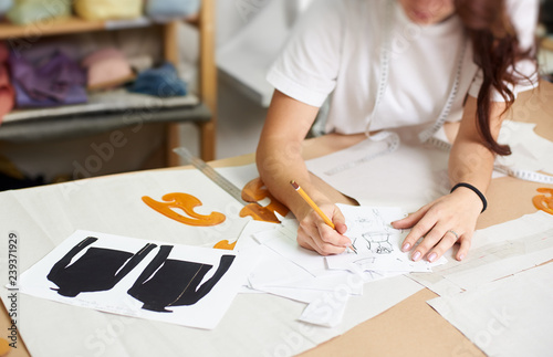 Fotografi Female designer drawing pencil sketches of clothing sitting at big table with flat paper patterns, measuring ruler and curves on blurred background of tailor design studio interior