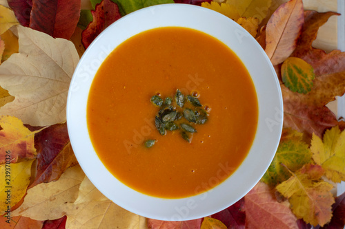 Tasty pumpkin bright orange soup with green seeds served on white plate and autumnal colorful leaves
