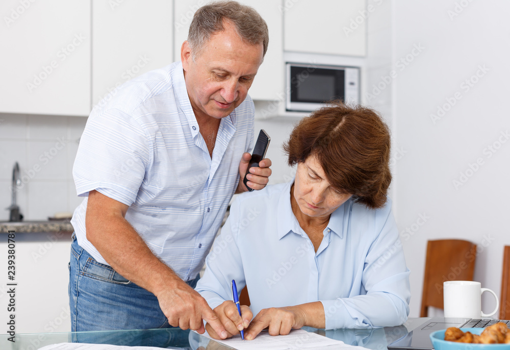 Fototapeta premium Mature couple at table in home kitchen, woman filling up documents