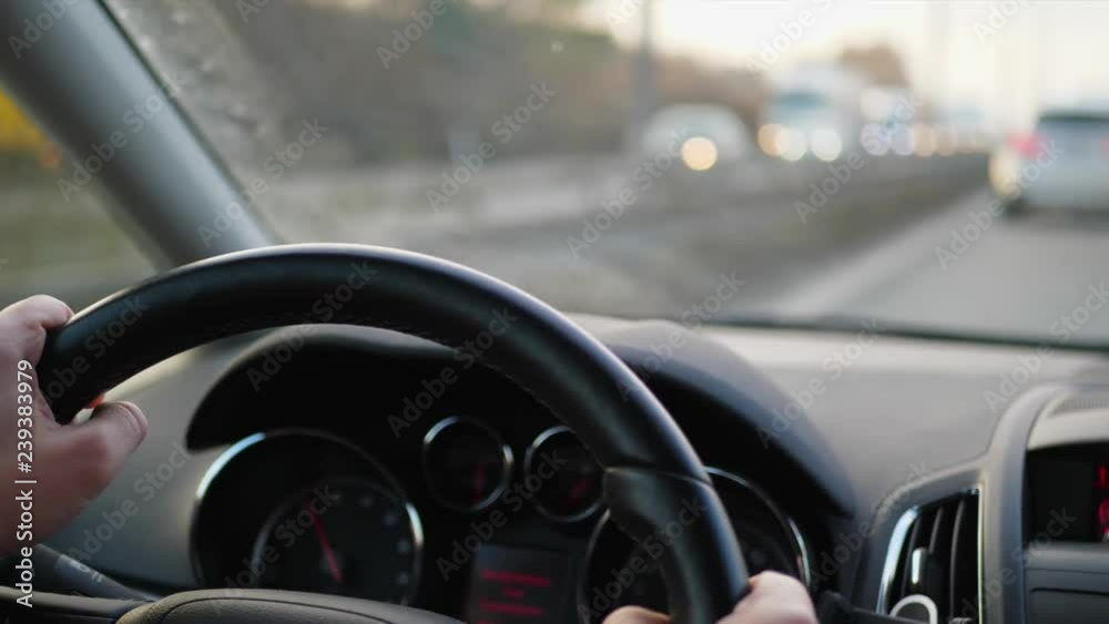 Close-up of steering wheel with hands on it during car driving. Transport drive on highway. Focus on wheel, sitter view inside of car. Unfocused transportation on active road seen in front window Close-up of steering wheel with hands on it during car driving. Transport drive on highway. Focus on wheel, sitter view inside of car. Unfocused transportation on active road seen in front window