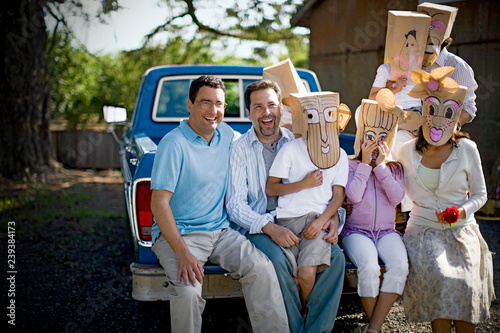 Portrait of two smiling mid-adult men sitting with their family who are wearing paper bag masks on the back of a truck. photo