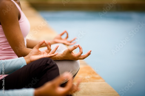 Two women sitting cross-legged with their thumbs and index fingers pressed together next to an outdoor swimming pool.