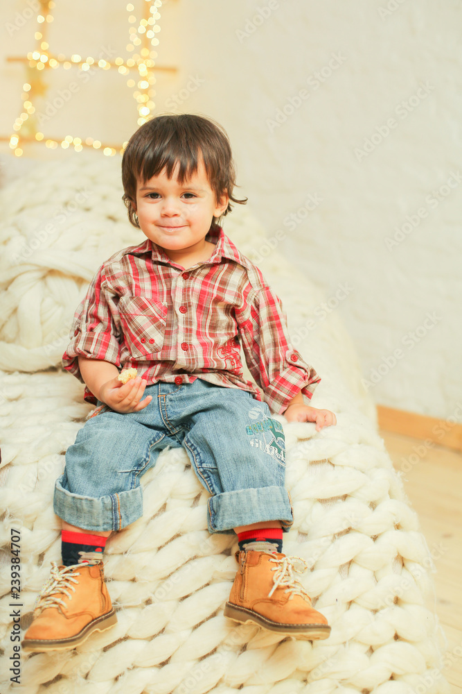 Nice cute small boy is sitting on knitted blanket on white bed, wearing checked shirt, jeans, leather boots, dark messy hair, smiling, eating bread Nice cute small boy is sitting on knitted blanket on white bed, wearing checked shirt, jeans, leather boots, dark messy hair, smiling, eating bread