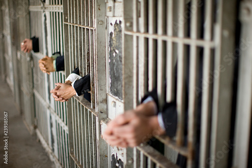 Hands of a mid-adult business woman and three colleagues through bars of a prison cell in a derelict building.