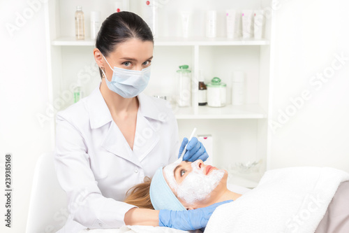 Relaxed young woman is getting facial skin care treatment at beauty salon. Beautician is touching brush with clay to face girl