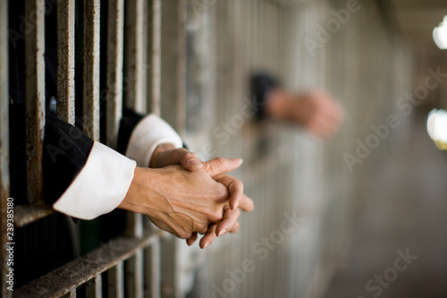 Hands of a mid-adult woman through bars of a prison cell in a derelict building.