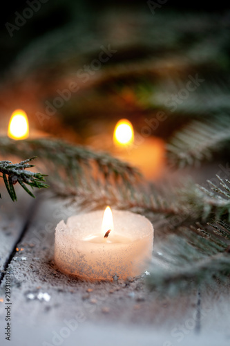 Burning candle and Christmas decoration over snow with pine branches on white wooden background.
