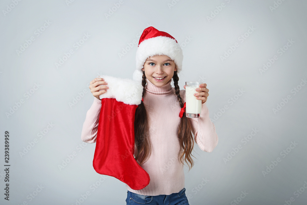 Young girl in santa hat holding christmas sock and glass of milk on grey background