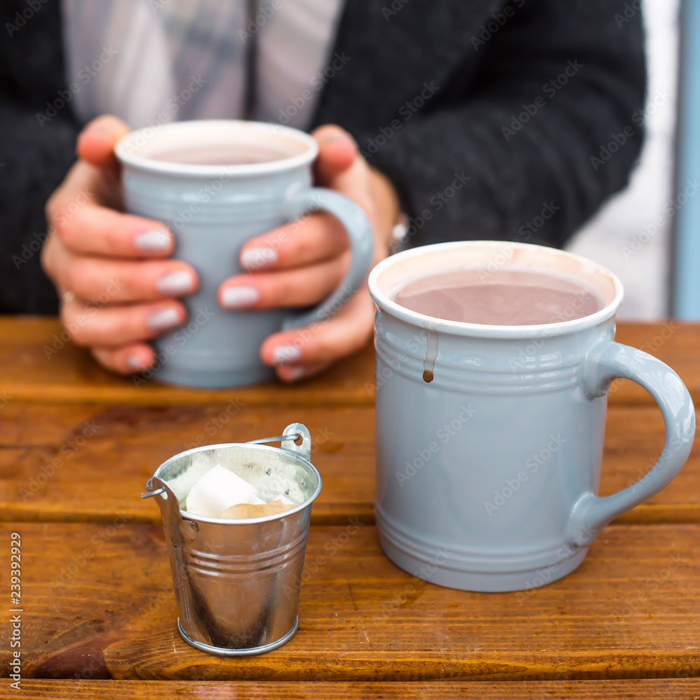 Φωτογραφία Two cups of cocoa on a wooden table, one of which is held in the hands of a woman in a warm coat