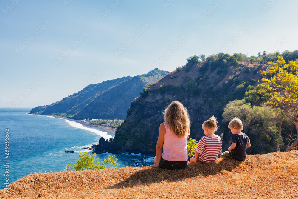 Family vacation lifestyle. Happy mother, kids on hill with scenic view of high cliffs, fishers village on black beach. Children looking at blue sea. Bukit Asah is popular travel destination in Bali.