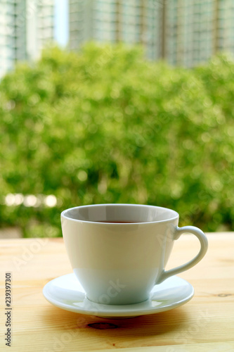 Vertical image of a cup of hot tea on the window side wooden table with blurred green foliage and high buildings in background 