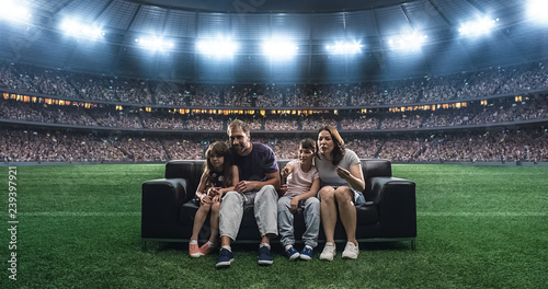 A family is watching a soccer moment, sitting on the couch located in the middle of the soccer stadium.