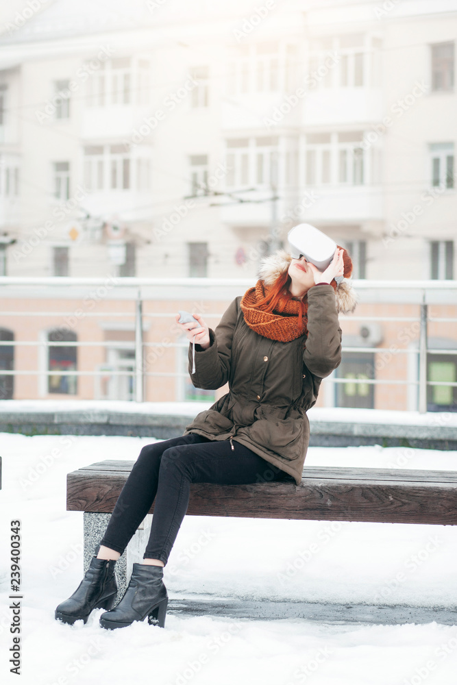 Pretty hipster young girl plays virtual reality mobile game app on smartphone attached to modern VR glasses outdoor in cold winter day Pretty hipster young girl plays virtual reality mobile game app on smartphone attached to modern VR glasses outdoor in cold winter day