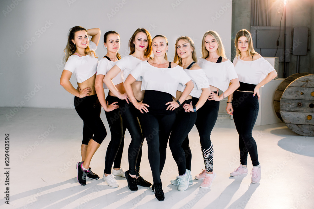 Group of young sporty girls in white and black outfit standing at white wall. Female companions in gym resting after fitness, indoor full length, loft studio, copy space