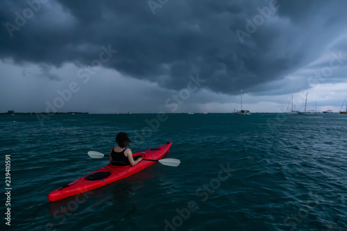 Adventurous girl on a red kayak is kayaking towards a thunderstorm during a dramatic sunset. Taken in Key West, Florida Keys, United States.