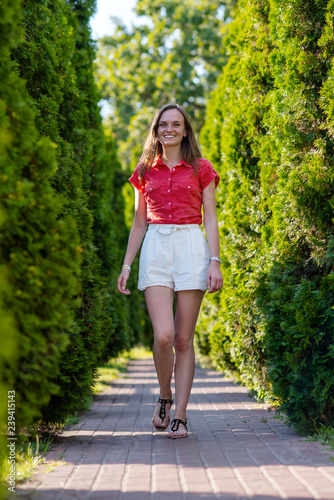 Portrait of young smiling woman in white short and red blouse walking in summer park, outdoors/Beautiful happy woman walking on the road