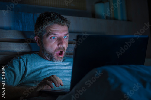 attractive internet addict man in shock and surprise networking late night on bed surprised face expression working with laptop computer in social media addiction