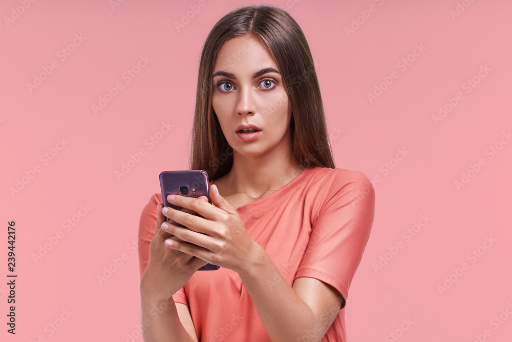 Beautiful female has attractive appealing appearance and shocked expression, received unexpected news on mobile phone, types message on modern electronic device, reads shocking promotional offer