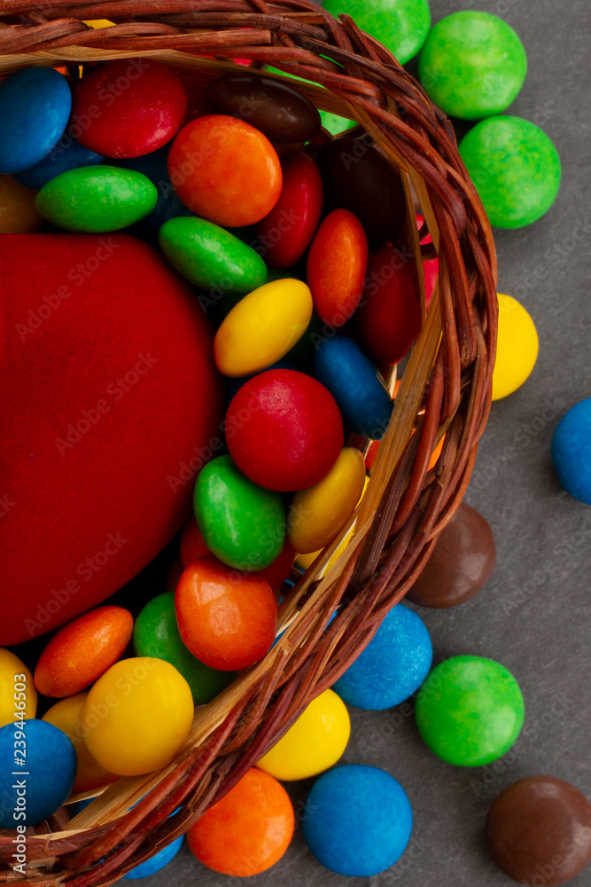 Portrait photo of a basket with sweets. Candy bowl with lots of  multicolored sweets and treats. A half of red velvet heart among a set of  bright glazed dragees. Stock Photo