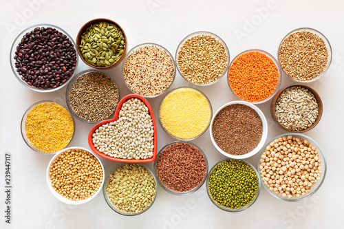 Top view to glass bowls with cereals, beans and seeds with red heart-shaped bowl in the middle on white background.