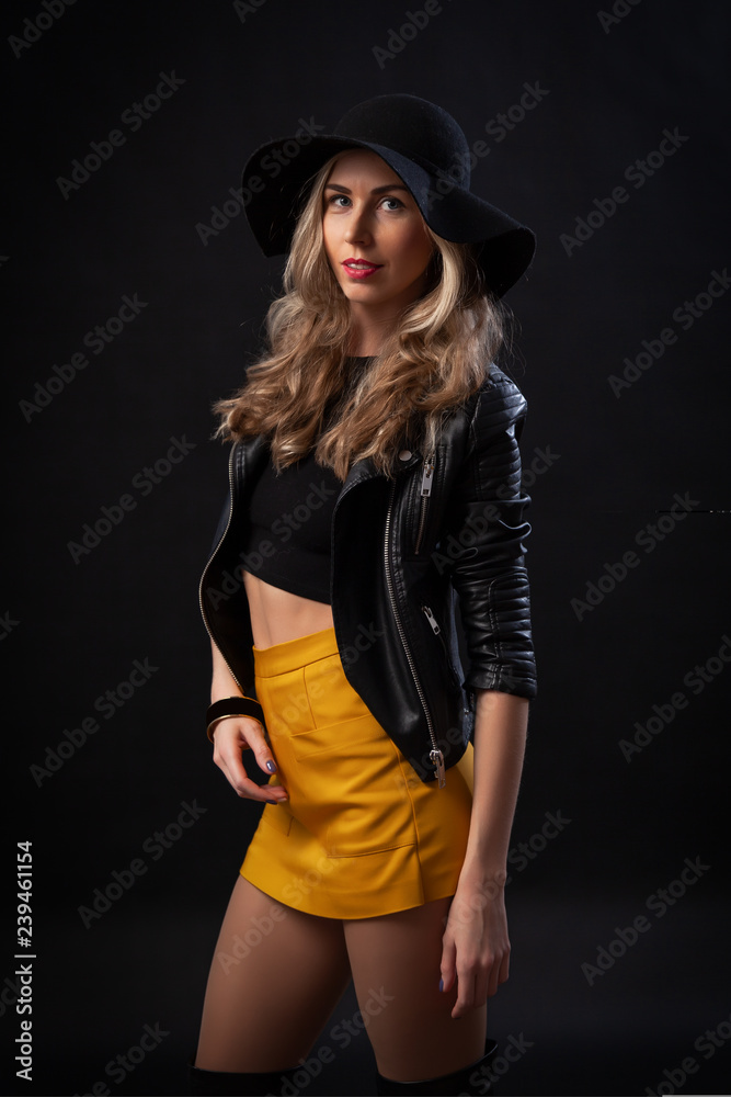 Portrait of wonderful young blonde woman with long hair in black T-shirt,  orange skirt, leather jacket and hat looking at camera and posing. Pretty  girl in trendy clothes. Curly hair Photos