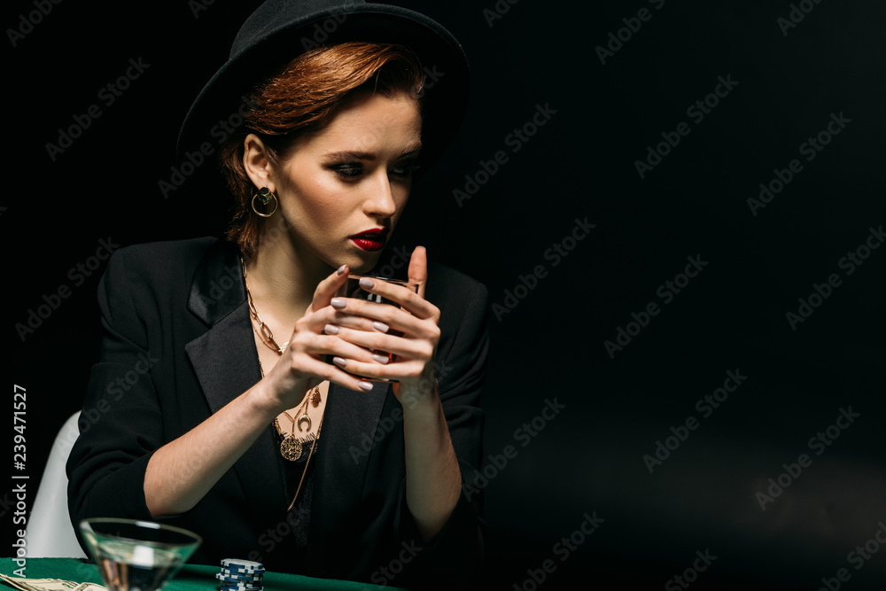 attractive girl in jacket and hat holding glass of whiskey at poker table in casino and looking away